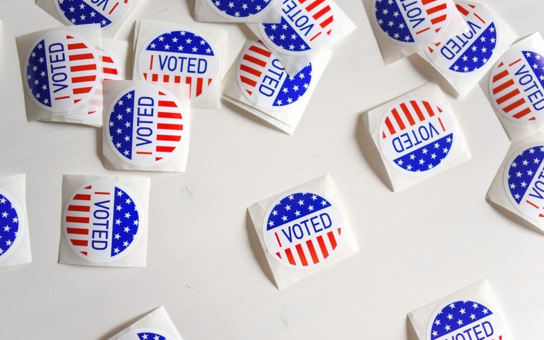 Circle shaped stickers whit a red, white and blue flag pattern on them that say "I Voted" strewn across a blank surface